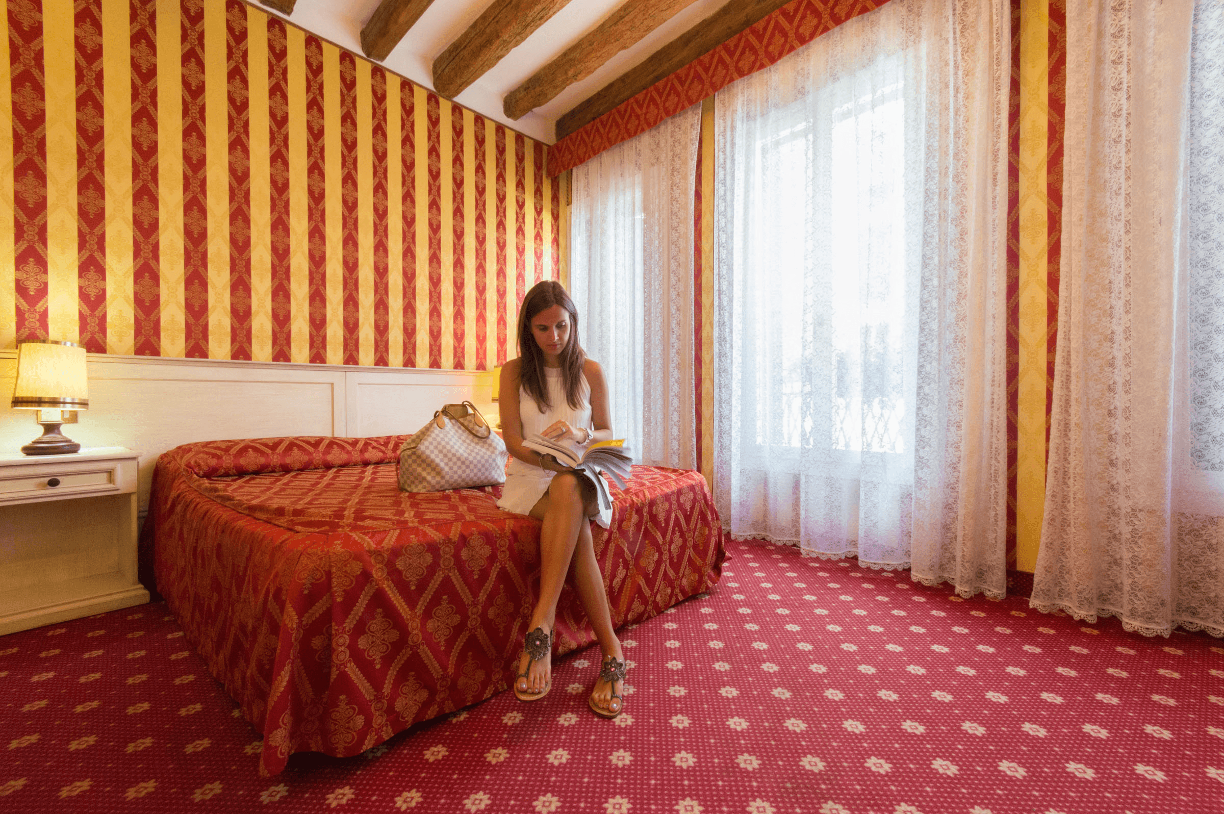 Donna in bianco legge su un letto rosso in una camera con pareti a righe.