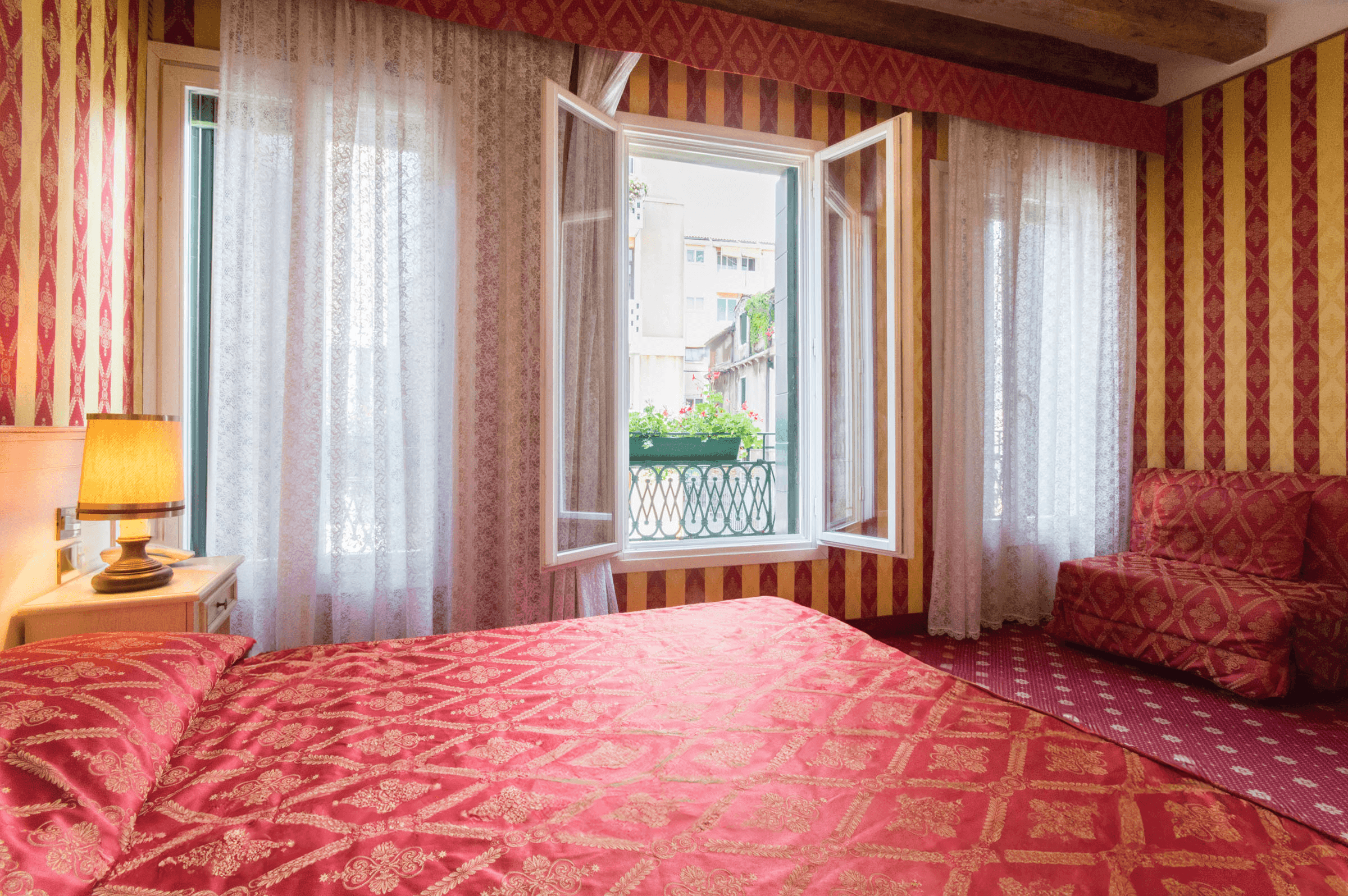 Camera da letto con pareti a righe rosse, copriletto damascato e finestra aperta sul balcone.