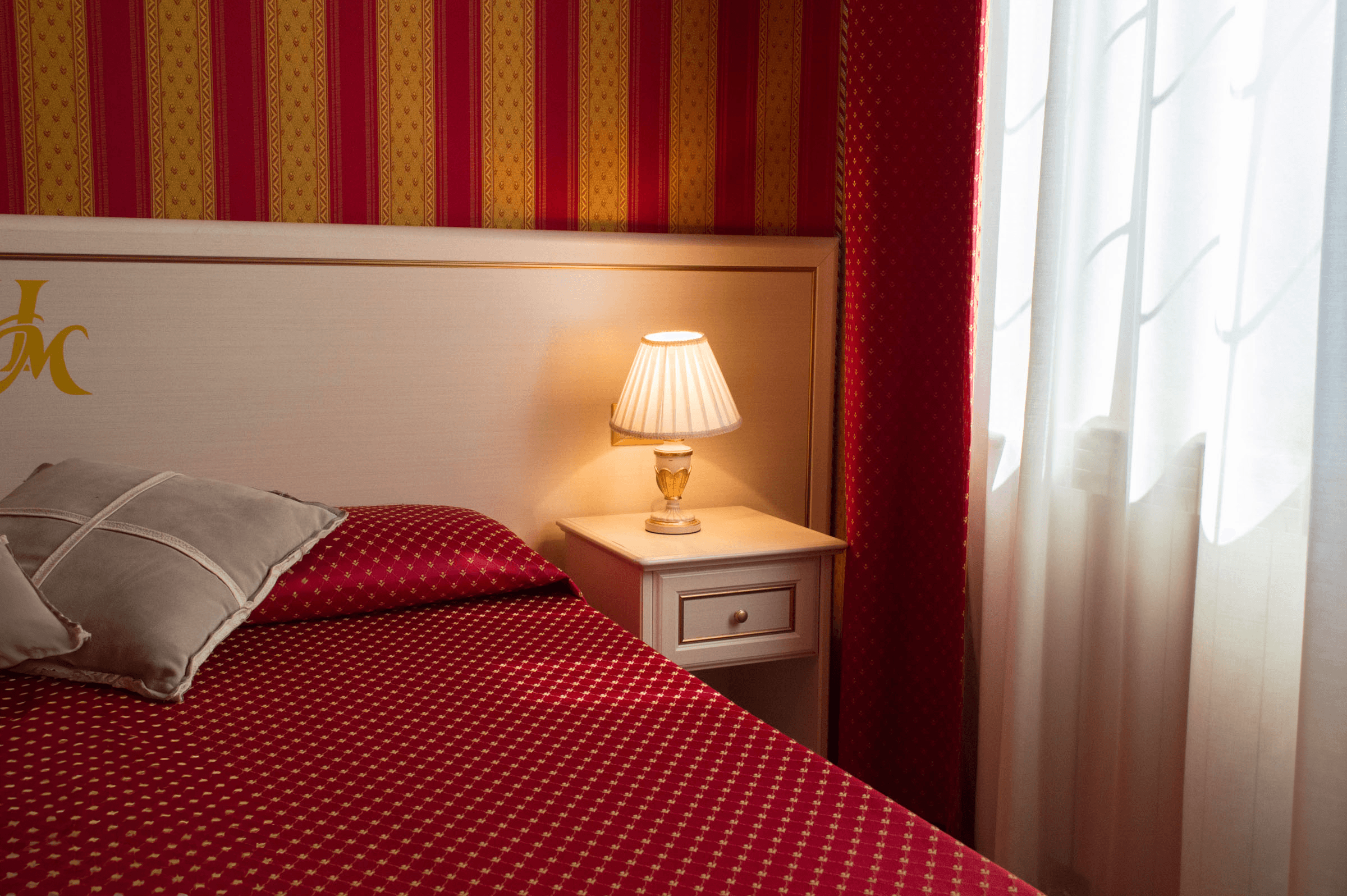 Letto con copriletto rosso, lampada accesa su comodino bianco e carta da parati a righe.