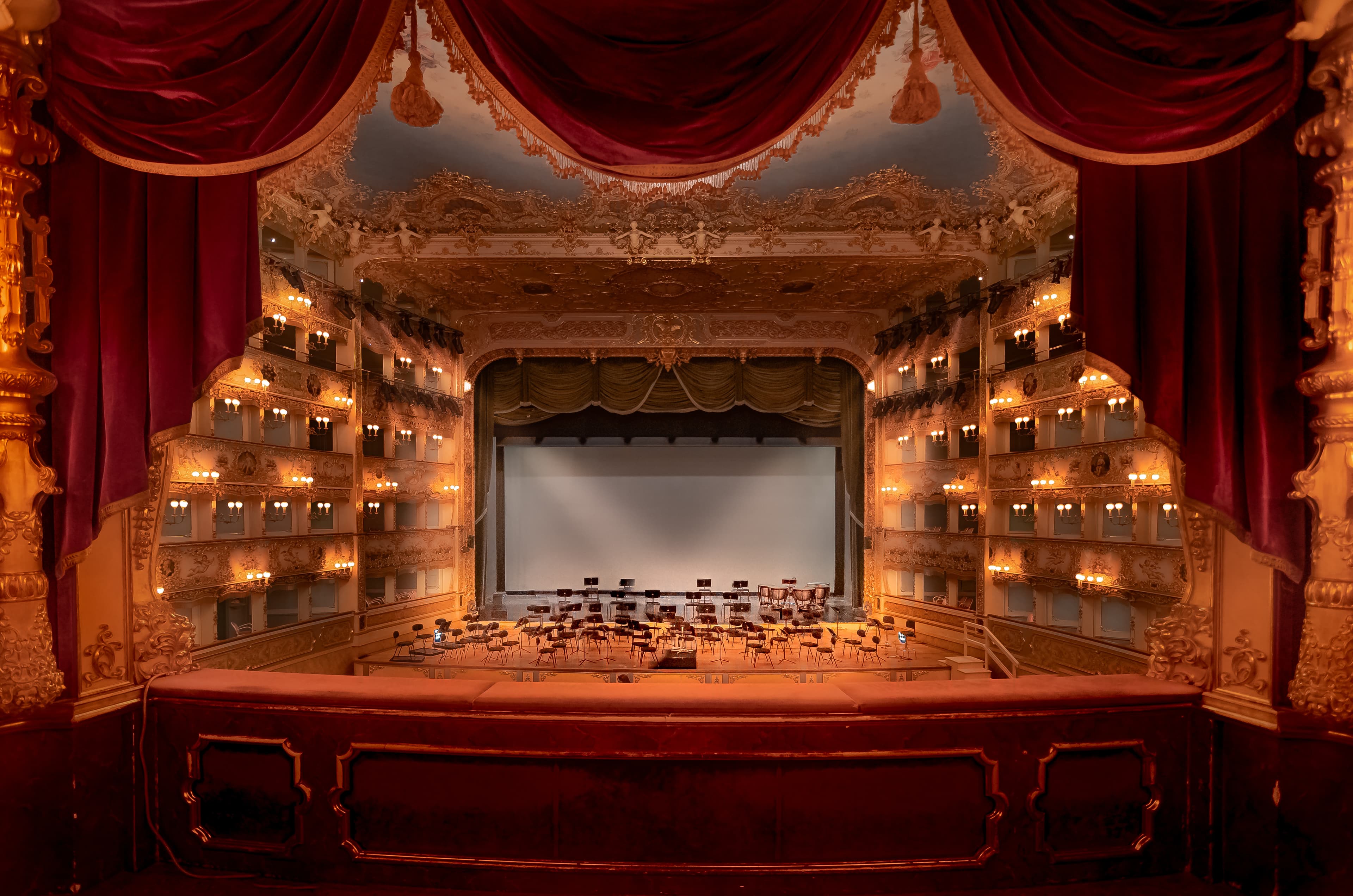 Teatro d'opera barocco con tende rosse, palchi dorati, sedie e leggii sul palco.